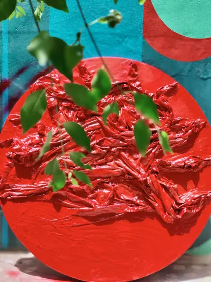 A textured, circular red artwork from the 'Reclaiming Red' series, seen here interacting with natural foliage.