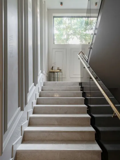This stairwell is designed to feel like a silent sculpture. It features clean marble treads, a paneled wall, and a glass railing with a brass handrail, all softly lit from a high window to create a moment of drama and pause.