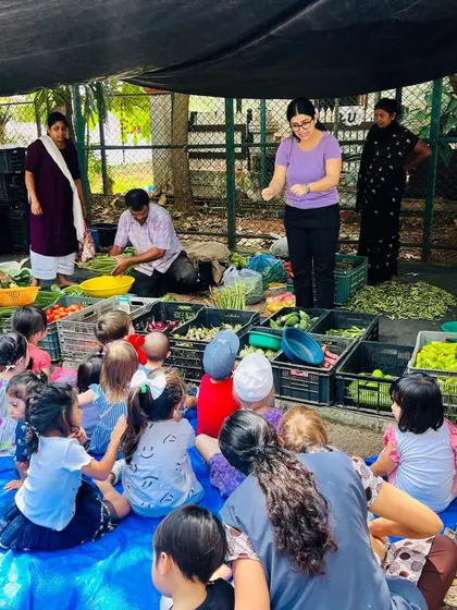 The children sit on a mat at the local market, listening as the vendor and teachers talk about the different vegetables.