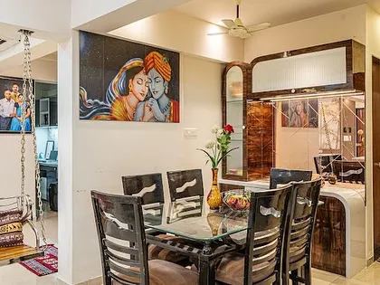 A classic dining setup with a glass-top table and a custom wooden crockery unit. The warm lighting and the Radha-Krishna painting create a welcoming atmosphere for family meals.