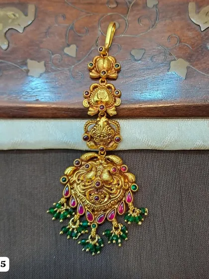 A beautiful 92.5 silver, gold-plated maang tikka featuring a traditional peacock design. The piece is embellished with red and green stones and green bead drops, perfect for a festive look.