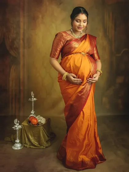 A serene solo portrait in a traditional orange saree. Cradling your bump, you look absolutely radiant against the classic studio backdrop with cultural props.