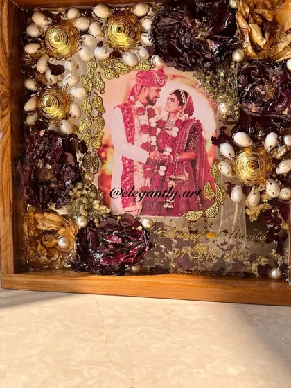 A close-up of a teakwood frame preserving kaleera and varmala flowers. The sunlight catches the details, making the piece glow with warmth and love.