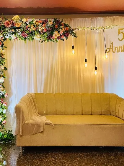 My decor style is versatile enough for any of life's great milestones. This elegant 50th-anniversary setup uses a sophisticated floral corner arch, soft drapery, and warm Edison bulb lighting to create a timeless and celebratory atmosphere.