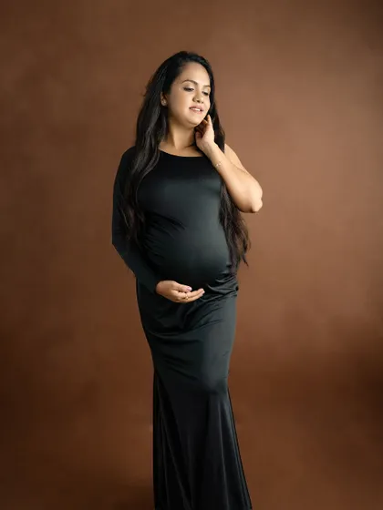 A simple yet stunning portrait of a mom-to-be in a black gown against a solid brown backdrop, emphasizing her form and serene expression.
