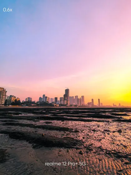 An ultra-wide 0.6x view of the Mumbai skyline at sunset. This mobile shot captures the vastness of the scene, from the foreground textures of the beach to the distant city lights.