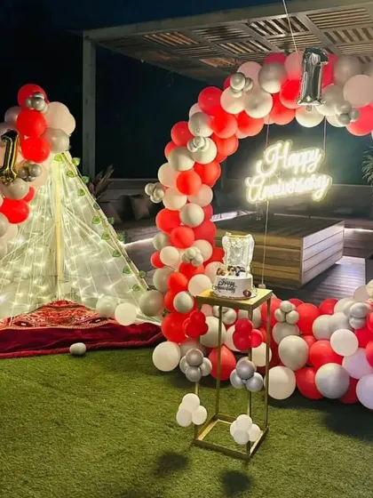 A first-anniversary party on a terrace with an artificial lawn. The setup features a circular balloon arch in red and white, a neon sign, and a cozy, lit-up teepee tent.
