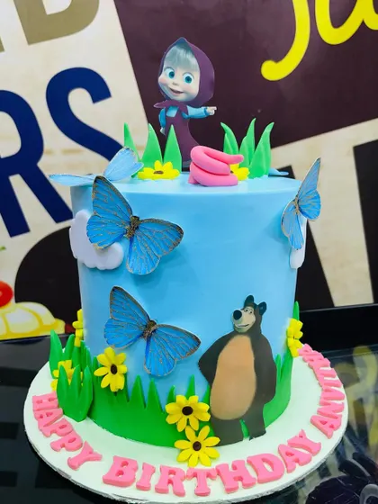 A playful 3kg Masha and the Bear cake set in a meadow with blue butterflies and yellow flowers.