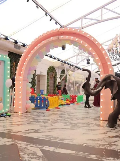 A grand entrance to a pastel carnival party, featuring a large rainbow arch, a light-up welcome sign, and a colorful play area, transforming an indoor venue into a magical space.