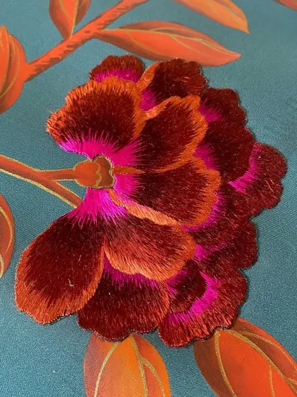 A close-up of an embroidered flower from the 'Folly' wallpaper. The rich, layered stitching in shades of red and pink on a teal silk background highlights the exquisite craftsmanship of the design.