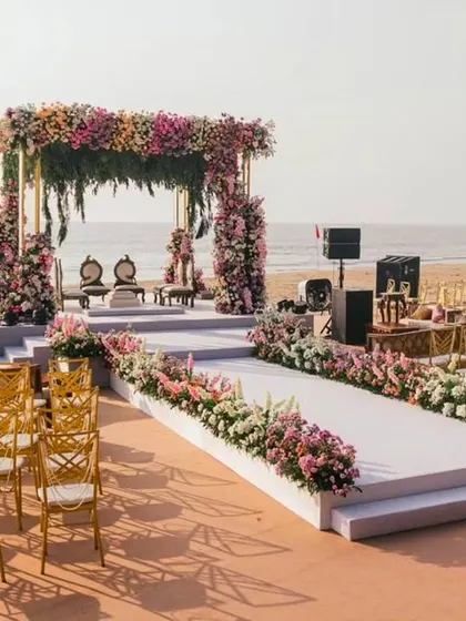 The elegant beach wedding mandap and aisle, ready for the ceremony. The combination of the elevated platform, the floral decor, and the golden chairs creates a stunning contrast against the sand and sea.