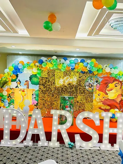 A bright and colorful jungle theme decoration with a gold shimmer wall. The stage features large marquee letters, a vibrant balloon arch, and Lion King character cutouts.