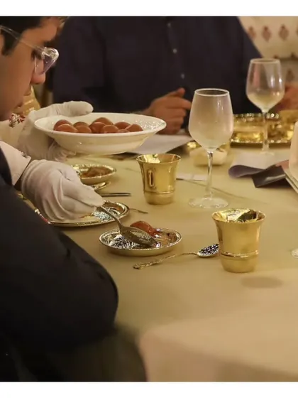 A close-up of dessert service at a formal dinner, where guests are served from a platter of traditional Indian sweets onto ornate silverware.