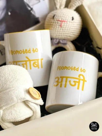 A close-up of the personalized mugs for 'Aaji' and 'Ajoba' in Marathi, paired with soft baby booties and a cute crocheted bunny rattle.