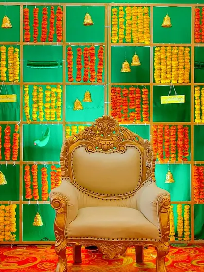 A close-up of a traditional stage setup. The backdrop is a grid of green panels filled with marigold flower strings and small brass bells, perfect for a Haldi or Sangeet ceremony.