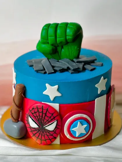 An epic Avengers-themed cake for a superhero fan. This design includes the Hulk's fist smashing through the top, Captain America's shield, Thor's hammer, and Spider-Man's mask, all handcrafted in fondant.