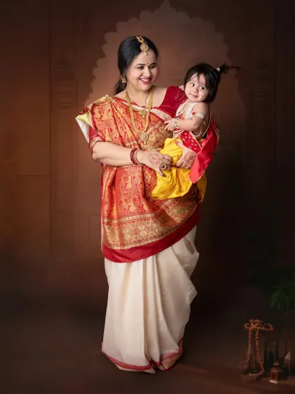 A mother dressed in a traditional saree holds her baby, who is costumed as little Krishna, creating a beautiful cultural portrait.