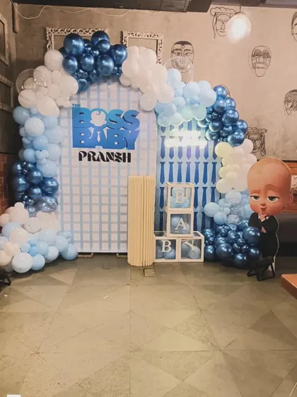 A fun "Boss Baby" themed decoration for Pransh. The setup includes a character cutout, a city-grid backdrop, and a blue and white balloon arch, perfect for celebrating a little boss.