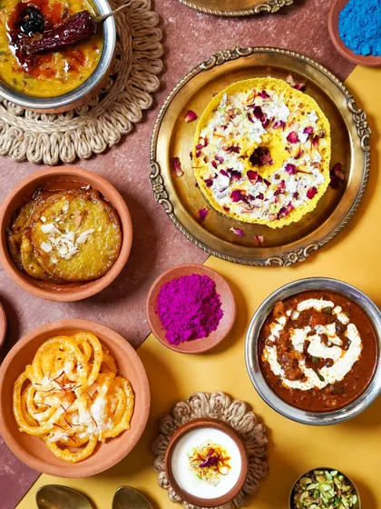 A vibrant spread of food for a Holi brunch, featuring both savory dishes and traditional sweets. The pops of color from the food and the scattered gulal capture the festival's joyful essence.
