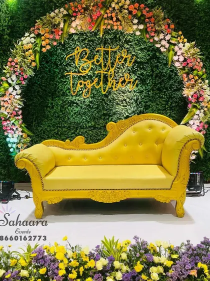 This vibrant photo booth features a bright yellow chaise lounge set against a lush green backdrop. The "Better Together" sign and a ring of colorful flowers make it a cheerful spot for pictures.