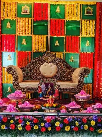 A vibrant and colorful Seemantham stage. The backdrop uses a grid of red, yellow, and green fabrics with floral accents, complemented by colorful offerings and large brass lamps.
