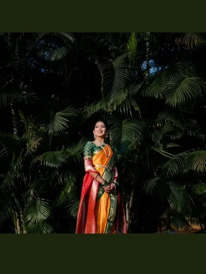 A stunning full-length portrait of the bride standing against a dark green, leafy backdrop, which makes her vibrant saree stand out.