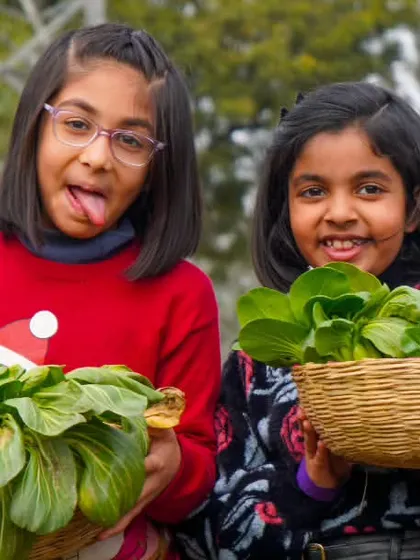 Two girls playfully show off their baskets filled with bok choy. Their fun and silly expressions capture the lighthearted atmosphere of the farm.