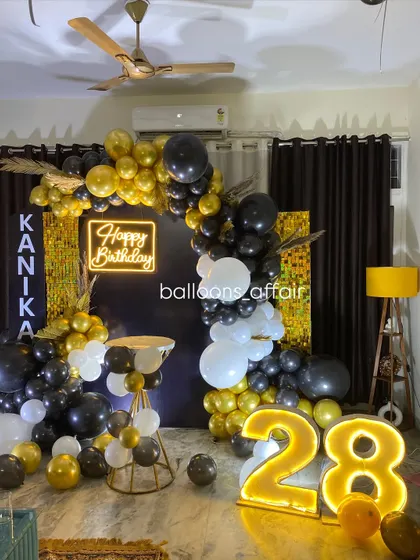 A chic 28th birthday party with a black and gold theme. The decor includes a balloon arch against a dark backdrop with a gold shimmer panel, a neon sign, and a light-up number.