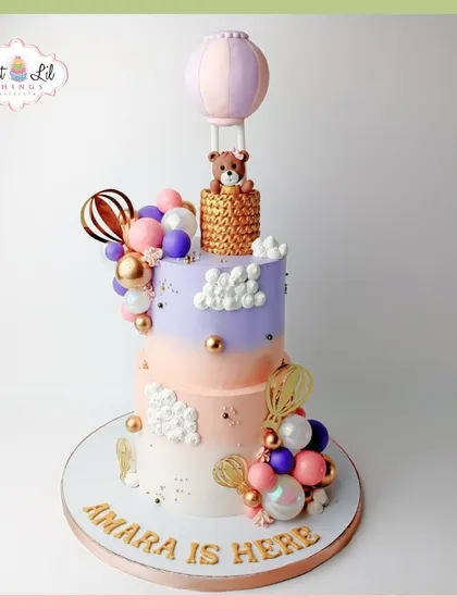 "Amara is here!" A dreamy welcome cake for a new baby, featuring a two-tiered design with hot air balloons, clouds, and a teddy bear.