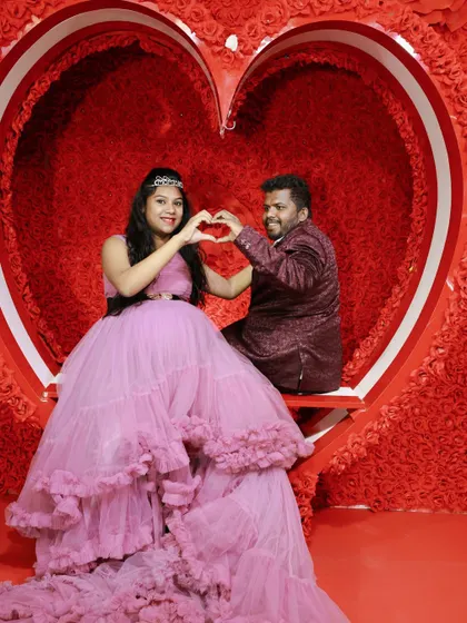 A happy couple making a heart shape with their hands during their maternity shoot. The mother-to-be is in a lavender gown, and they are seated in our heart prop.