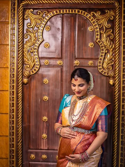 A portrait framed by a grand, ornate doorway. The traditional architecture complements the elegance of the saree and jewelry, creating a regal and timeless maternity photograph.