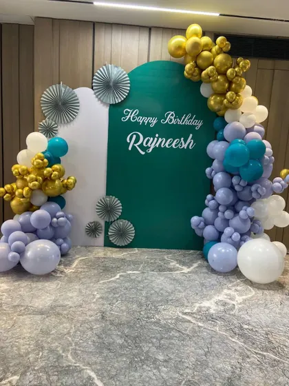 A sophisticated and personalized 50th birthday backdrop. We used custom arched panels in white and deep green, adorned with an organic balloon garland in lavender, teal, and chrome gold for a unique and modern celebration.