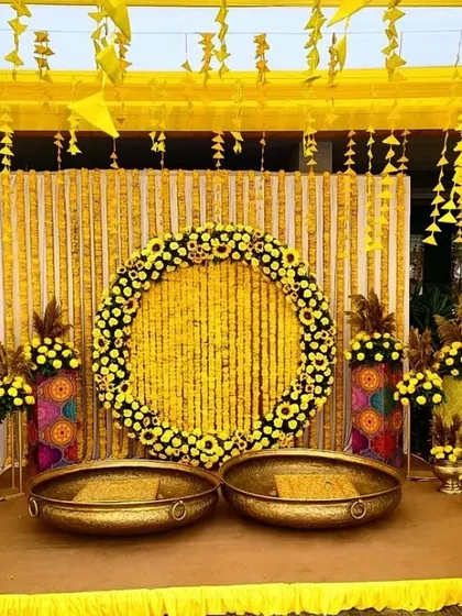 A classic Haldi stage decorated entirely in yellow. The centerpiece is a large wreath made of marigolds, surrounded by traditional brass urlis.