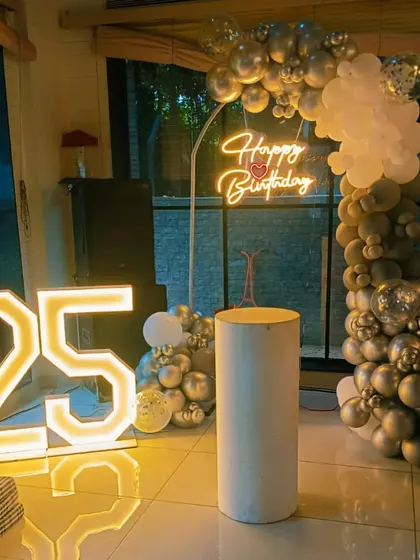 A 25th birthday party with a silver and white balloon arch, a 'Happy Birthday' neon sign, and a large '25' marquee light.