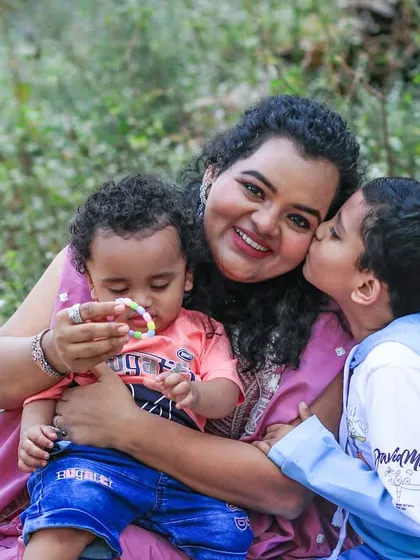 A mother embraces her two sons in a grassy field, with the older boy giving her a sweet kiss on the cheek. This is a heartwarming portrait of maternal love and sibling affection.