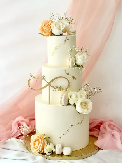 An elegant three-tier cake with a simple off-white buttercream finish. It's decorated with a scattering of gold leaf, fresh baby's breath, and clusters of peach and white roses.