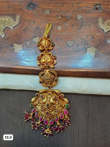 This traditional maang tikka features a detailed peacock design and is crafted from 92.5 silver with gold plating. The ruby-colored bead drops make it perfect for bridal wear.