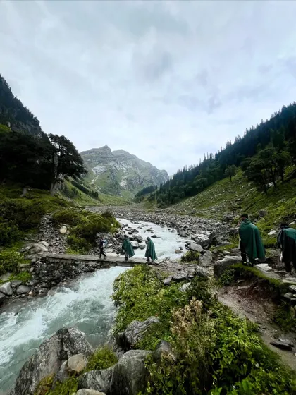 Trekkers in ponchos crossing a river on a rustic wooden bridge. Rain or shine, the adventure on the Hampta Pass never stops.