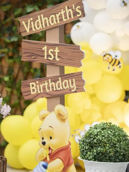 A '1st Birthday' sign for Vidharth's Pooh-themed party. The wooden sign and Pooh cutout perfectly match the rustic, gentle theme of the celebration.