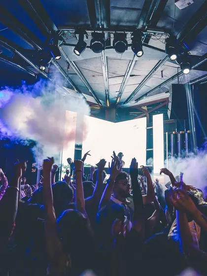 A powerful shot of smoke cannons erupting in front of the DJ booth, with the crowd's hands raised in excitement.