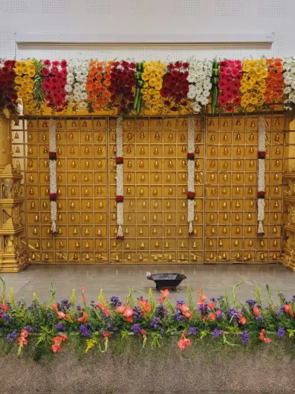 A grand stage setup with a golden backdrop of bells, topped with a thick, multi-colored floral canopy. The foreground is lined with a bed of fresh flowers.