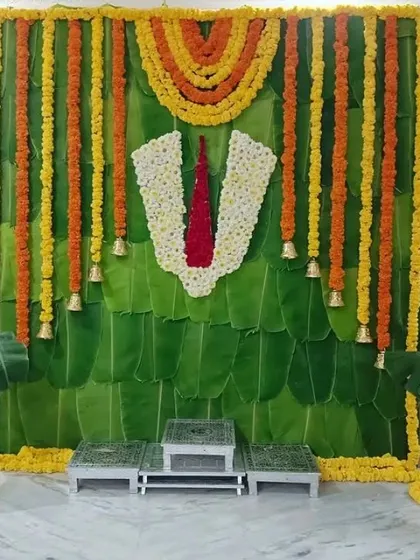 A South Indian style decoration featuring a backdrop of banana leaves, marigold garlands, and a central floral motif of Lord Balaji's tilak.