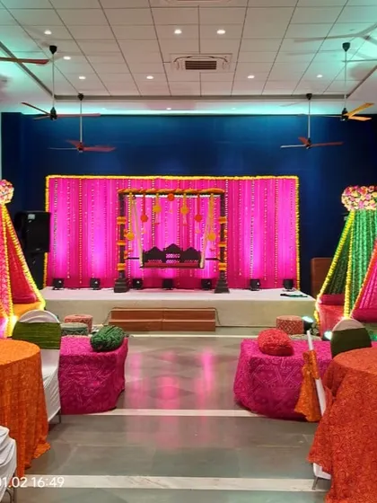 A colorful Mehendi setup with bright drapes in orange, pink, and green. The decor includes a traditional jhoola on the stage and low seating with bandhani print cushions for a festive feel.