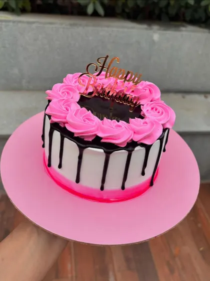 A pretty pink and black drip cake. The design features a dark chocolate drip over white buttercream, topped with pink buttercream rosettes.