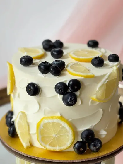 A refreshing lemon and blueberry cake. The white buttercream has a rustic texture and is decorated with fresh blueberries and slices of lemon.