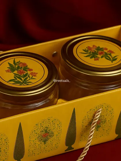 A classic and elegant Diwali gift. This set contains two glass jars with traditional floral-patterned lids, presented in a matching yellow gift bag, ready to be filled with festive treats.