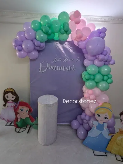 A sweet princess-themed naming ceremony backdrop. The personalized sign 'Hello World I'm Dhanasvi' is surrounded by a pastel balloon arch and cutouts of beloved Disney princesses.