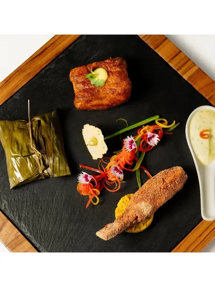 An assorted platter of appetizers, including a banana leaf-wrapped item and a fried fish fillet. The dish is artfully arranged on a slate board, showcasing the chef's plating for a fine-dining menu.