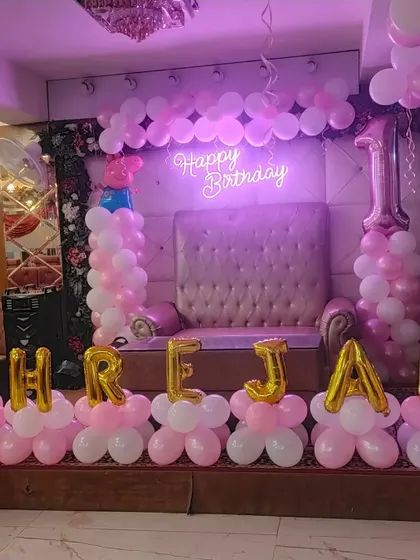 A first birthday stage setup in pink and white, with a large throne-like chair for the birthday girl and her name in foil letters.