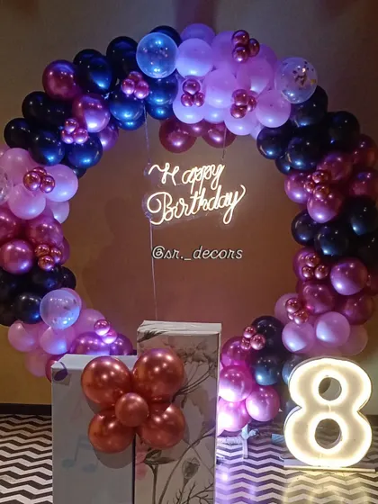 A stylish 8th birthday decor using a ring of dark blue, pink, and black balloons, a neon sign, and a light-up number.